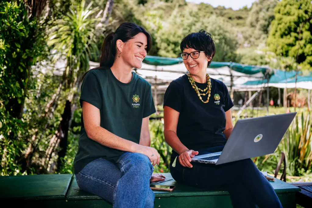 Two women in branded t-shirts smiling and chatting while using a laptop outdoors.