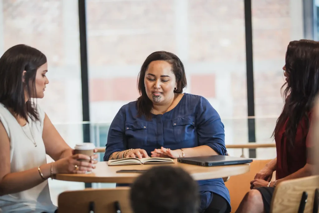 Three women sitting around a table, engaged in a meeting. One woman in a blue top is reading from a notebook while the others listen.