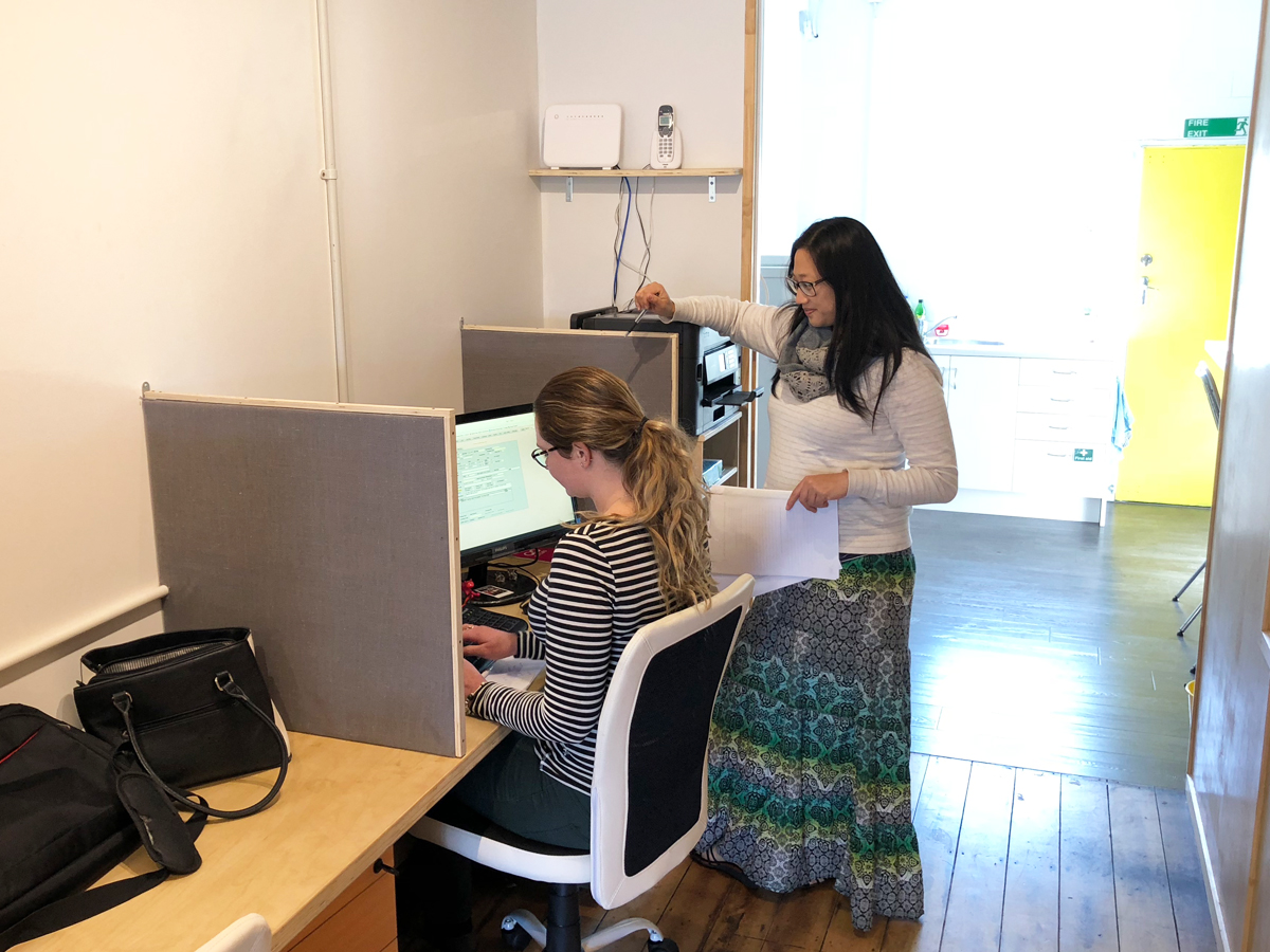One person works at a computer in a coworking space while another person stands next to them chatting