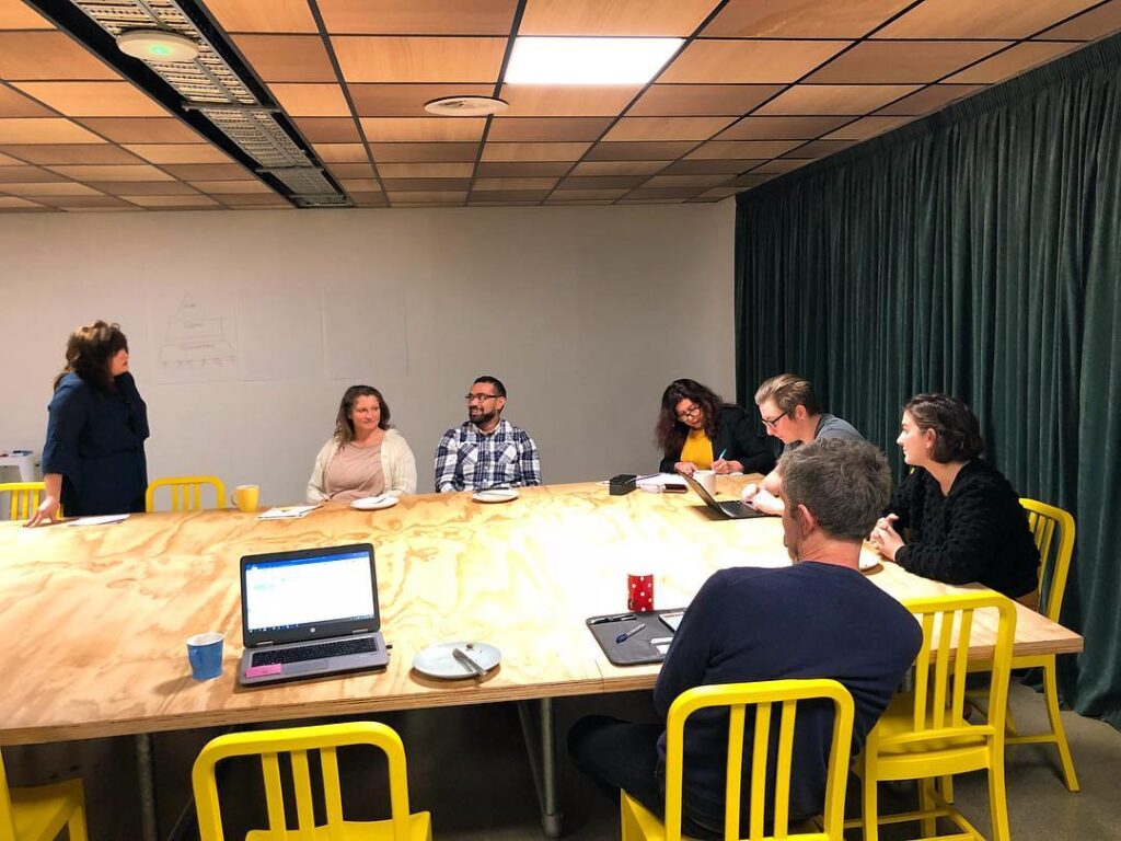 People sit around a wooden table in a meeting room, engaged in discussion while one woman stands and addresses the group.