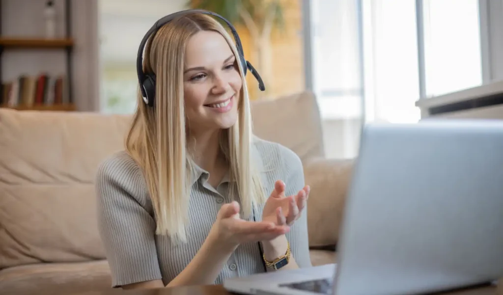 A woman with a headset smiles and gestures while video chatting on a laptop. She is seated on a beige sofa in a well-lit room, creating a warm, friendly tone.