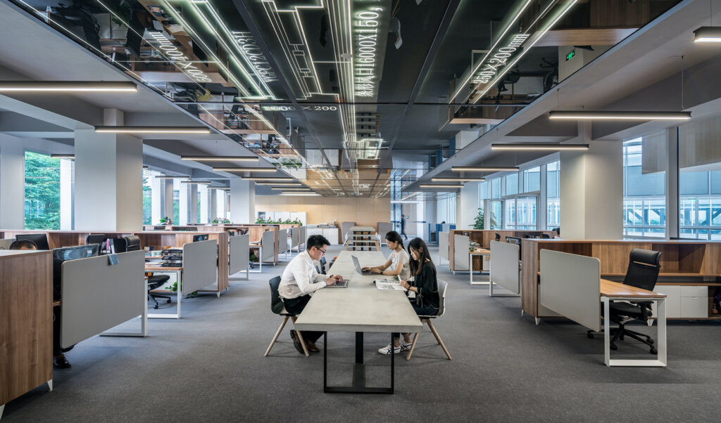 Modern open-plan office with employees working at desks and a central table, featuring bright lighting, large windows, and reflective ceiling panels.