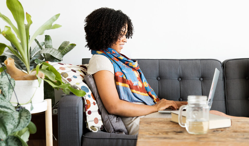 Woman working on a laptop from home, seated on a couch with plants, books, and a drink on the table.