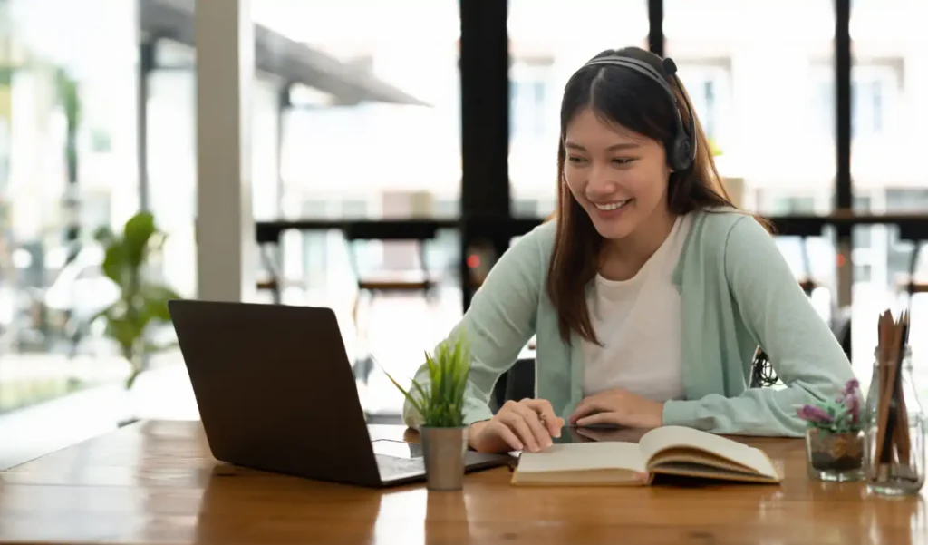 Smiling woman with headphones sits at a wooden table, engaging with a laptop. Open notebook and plants nearby, suggesting a relaxed work environment.