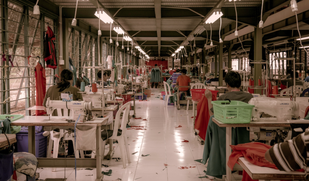 Workers sit at sewing machines in a large garment factory, surrounded by fabric, thread, and clothing under fluorescent lights.