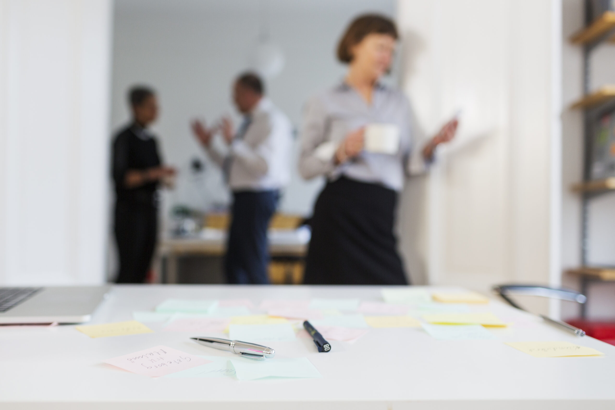 A desk in the foreground with pens and sticky notes scattered, while business professionals in the background engage in a conversation, partially out of focus