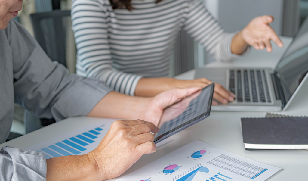 Two people sit at a desk, discussing something. Their torsos and arms are only visible - one has a laptop, the other a tablet