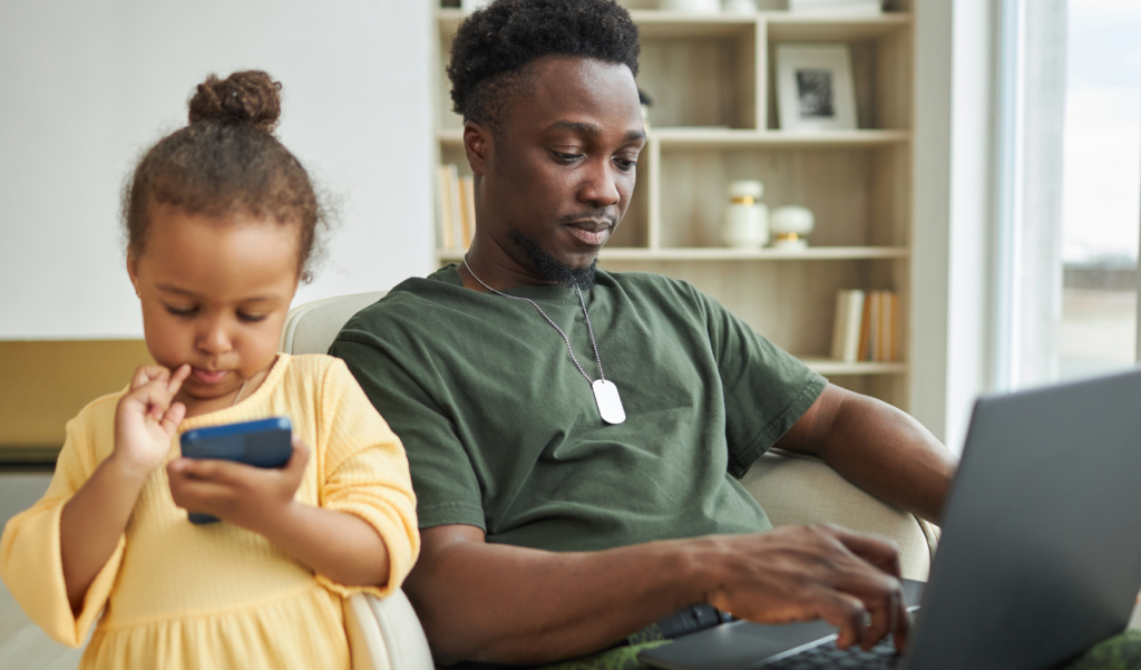 Man working on a laptop at home while a young girl beside him looks at a smartphone, both in a cozy, well-lit living space.
