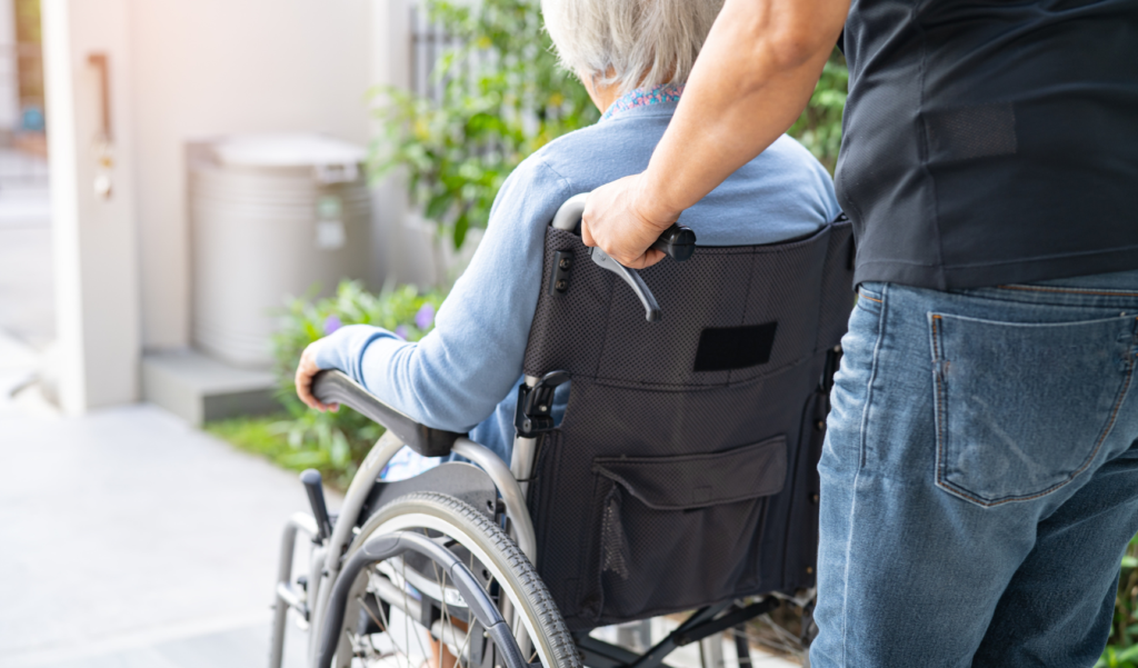 Caregiver pushing an elderly person in a wheelchair outdoors near a home entrance, with plants and sunlight in the background.