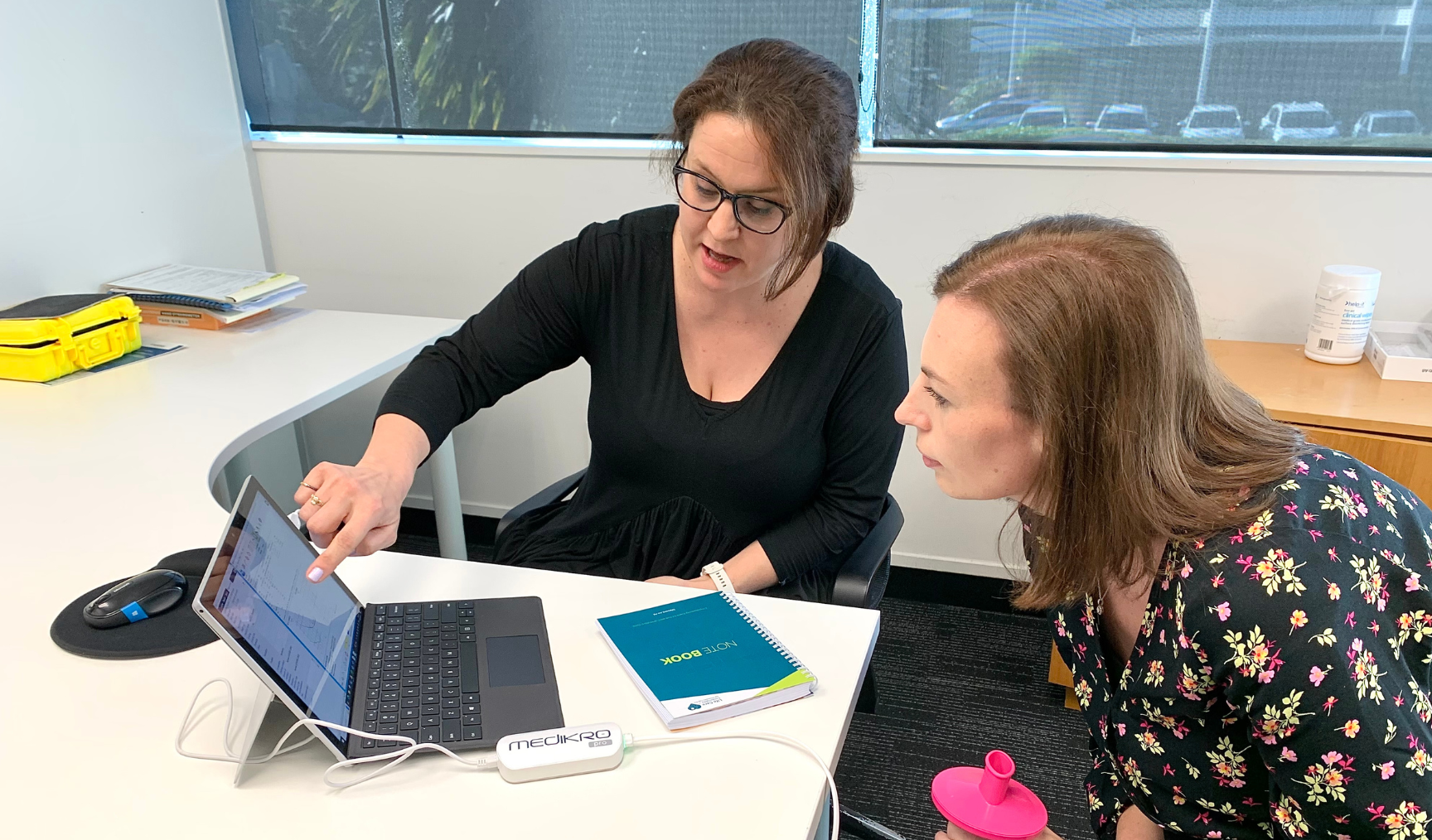 Two women are seated in an office, one pointing at a laptop screen while the other leans in to observe