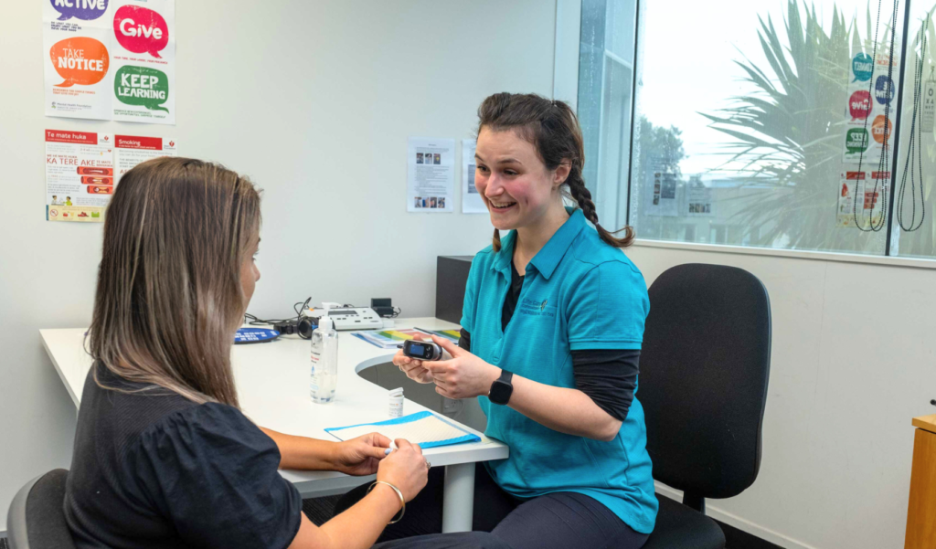 Smiling woman in a teal shirt sitting at a table, showing something on a device to another woman seated across from her. Posters and a window are visible in the background.