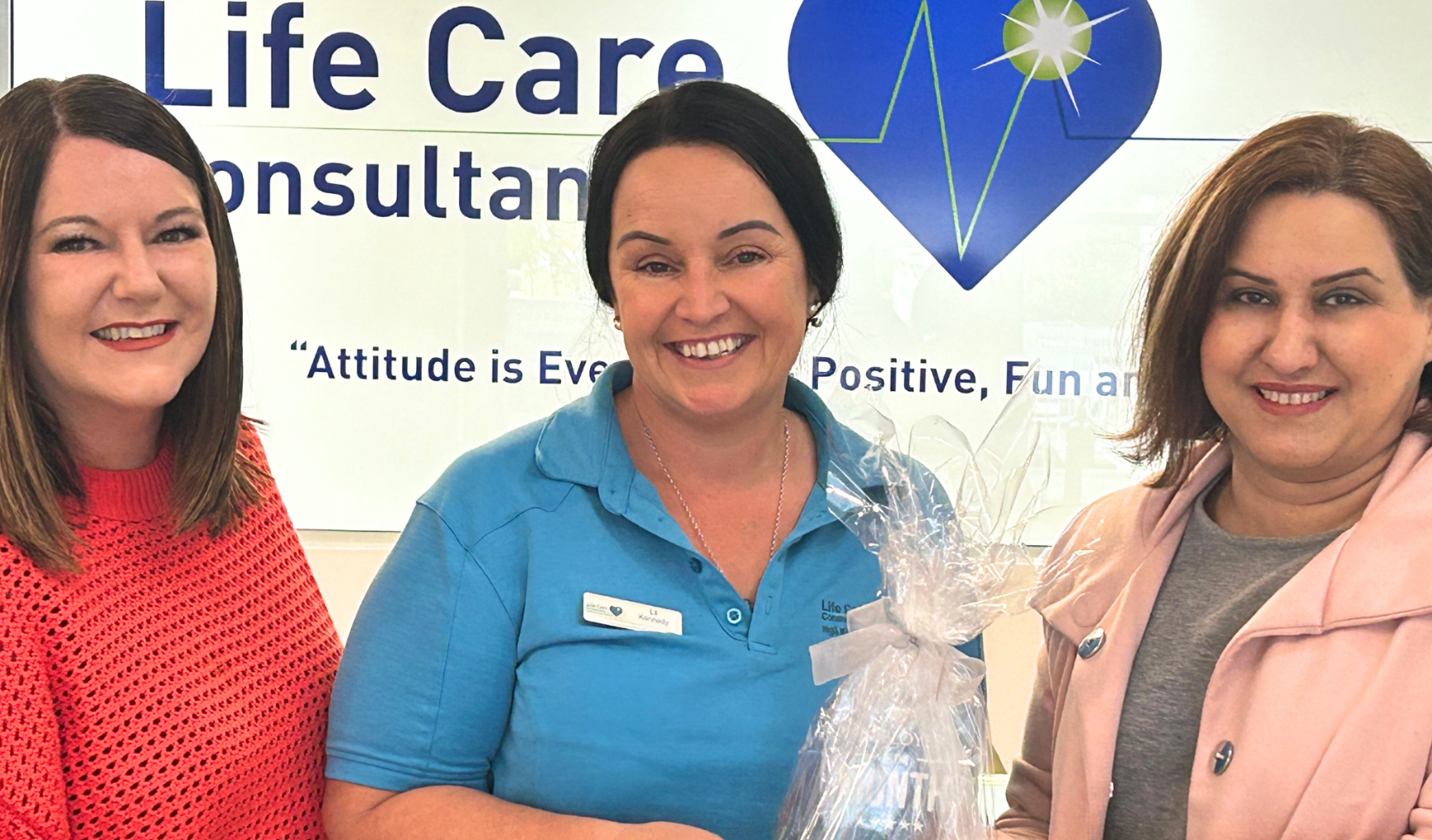 Three women pose for a picture, smiling with Life Care logo in the background. The woman in the middle holds a wrapped reward