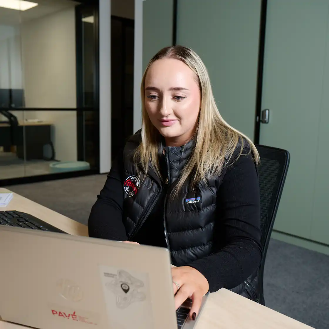 Woman with long blonde hair working on a laptop in a modern office, wearing a black vest with badges. She appears focused and engaged.