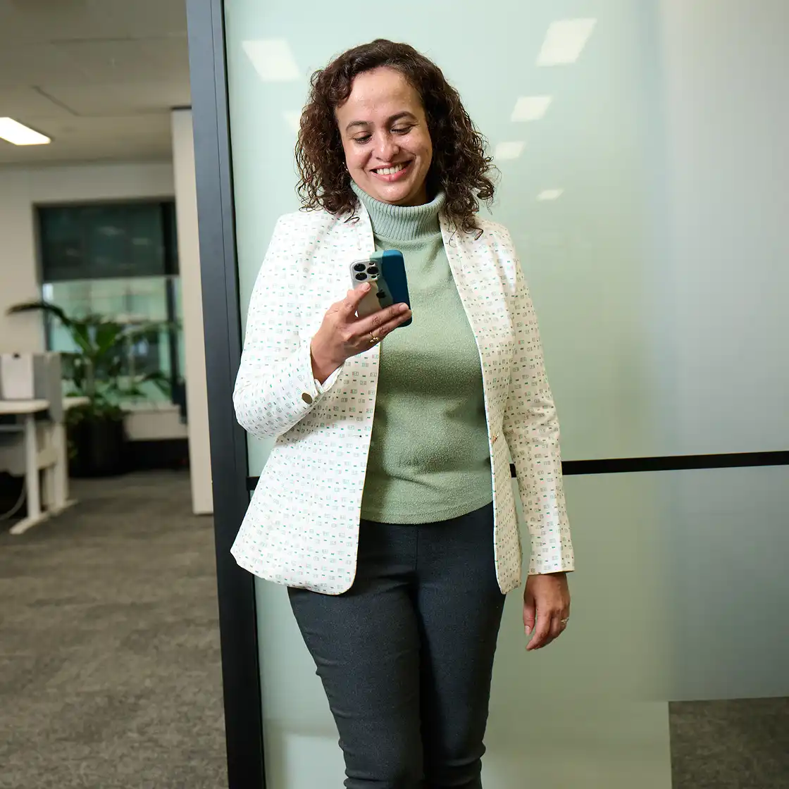 Smiling woman in a white blazer and green turtleneck looks at her phone in a modern office. She stands by a frosted glass wall, conveying a positive mood.