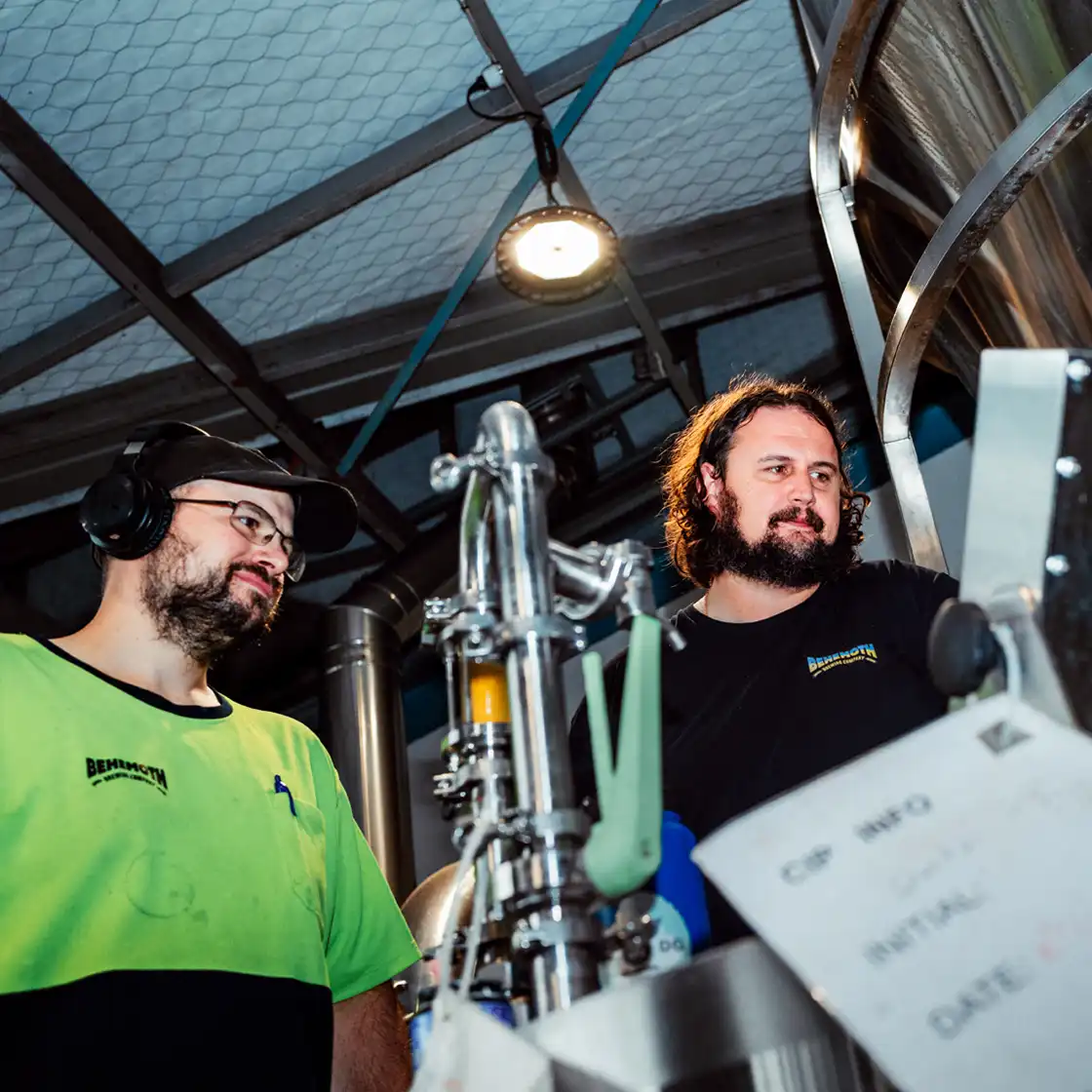 Two men in a brewery examine stainless steel equipment under bright lighting. One wears ear protection, creating a focused and industrious atmosphere.