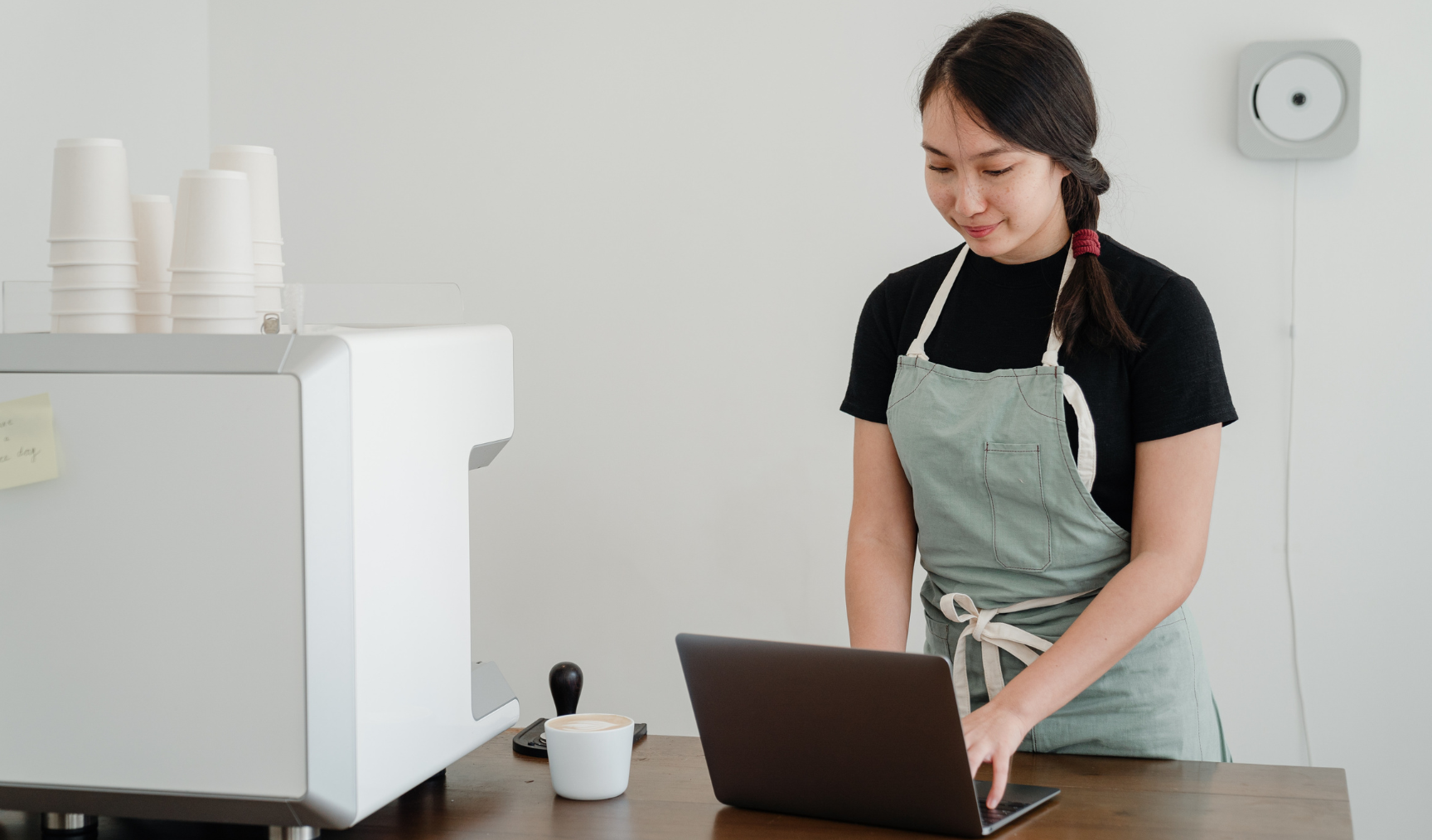 A woman works on her laptop on a table with a barista coffee machine