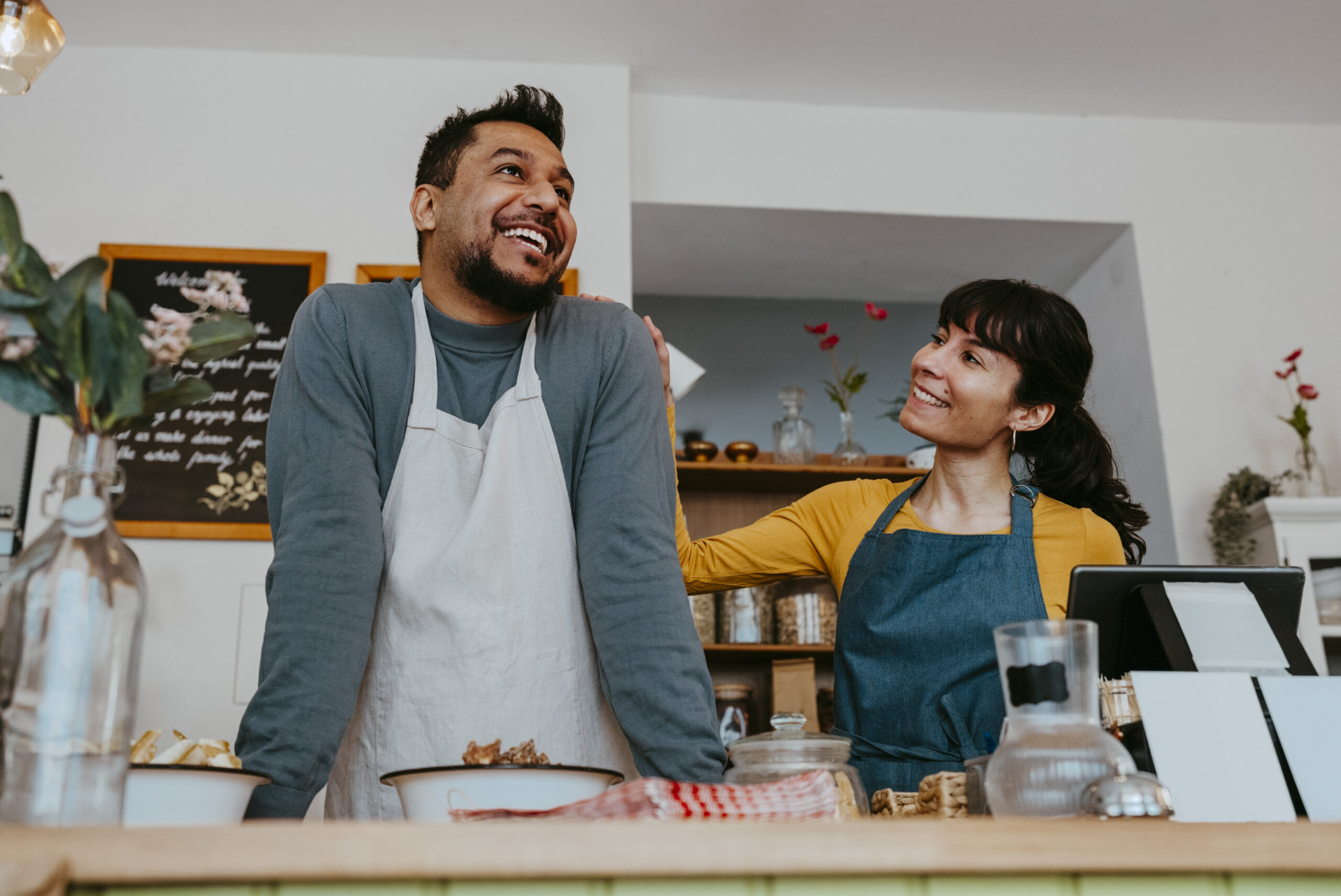 Two small business owners, a man and woman in aprons, smile and share a light moment behind the counter of a cozy shop or café.
