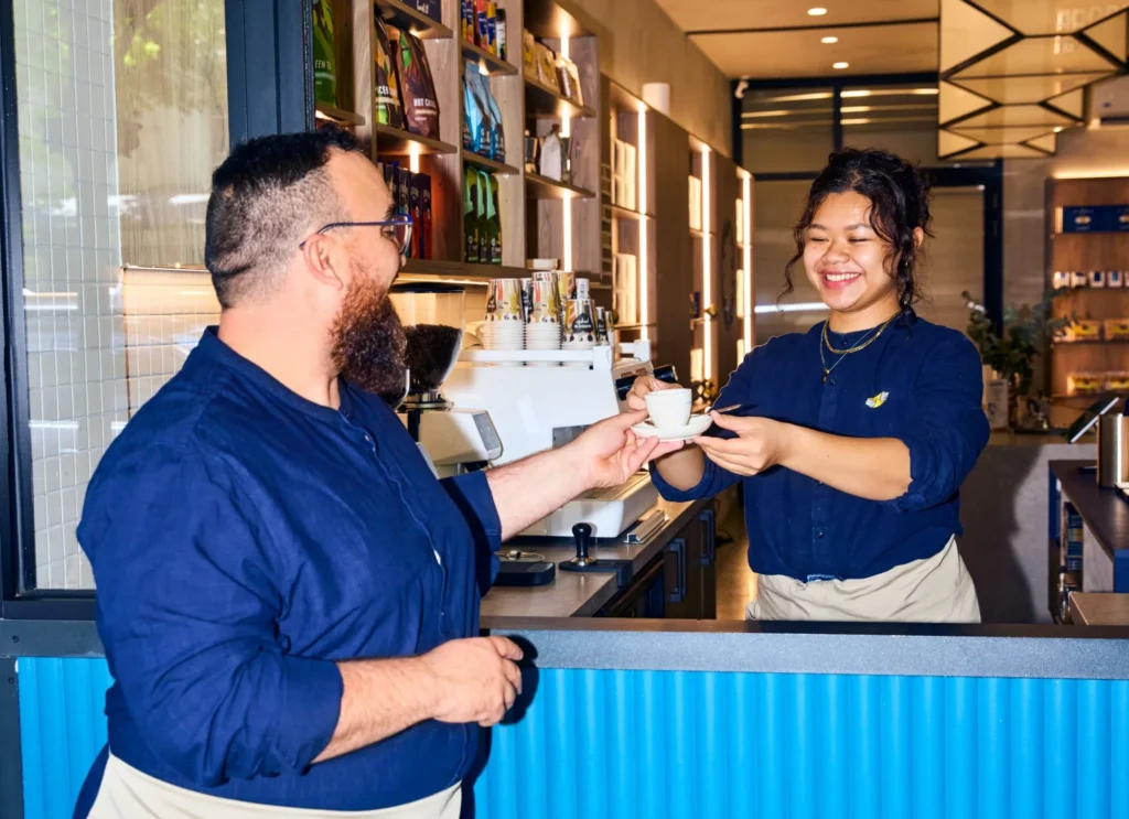 Barista smiling as she hands a cup of coffee to a customer across the café counter.