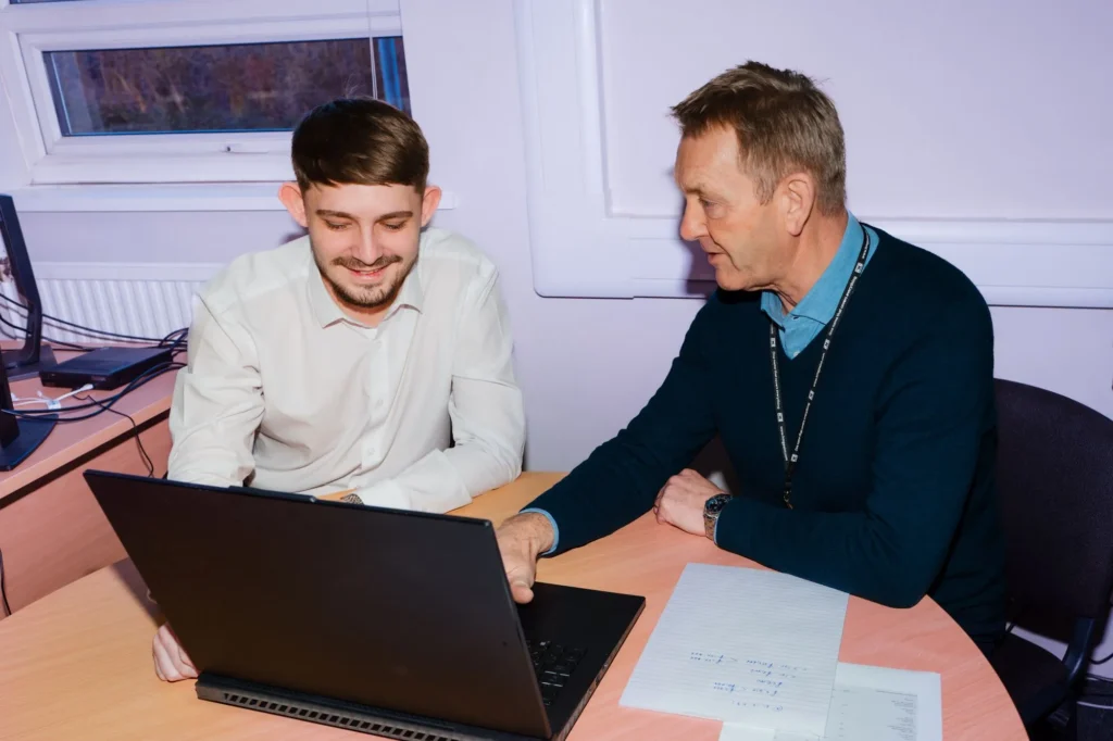 Two men sitting at a desk, smiling while looking at a laptop during a collaborative work session.