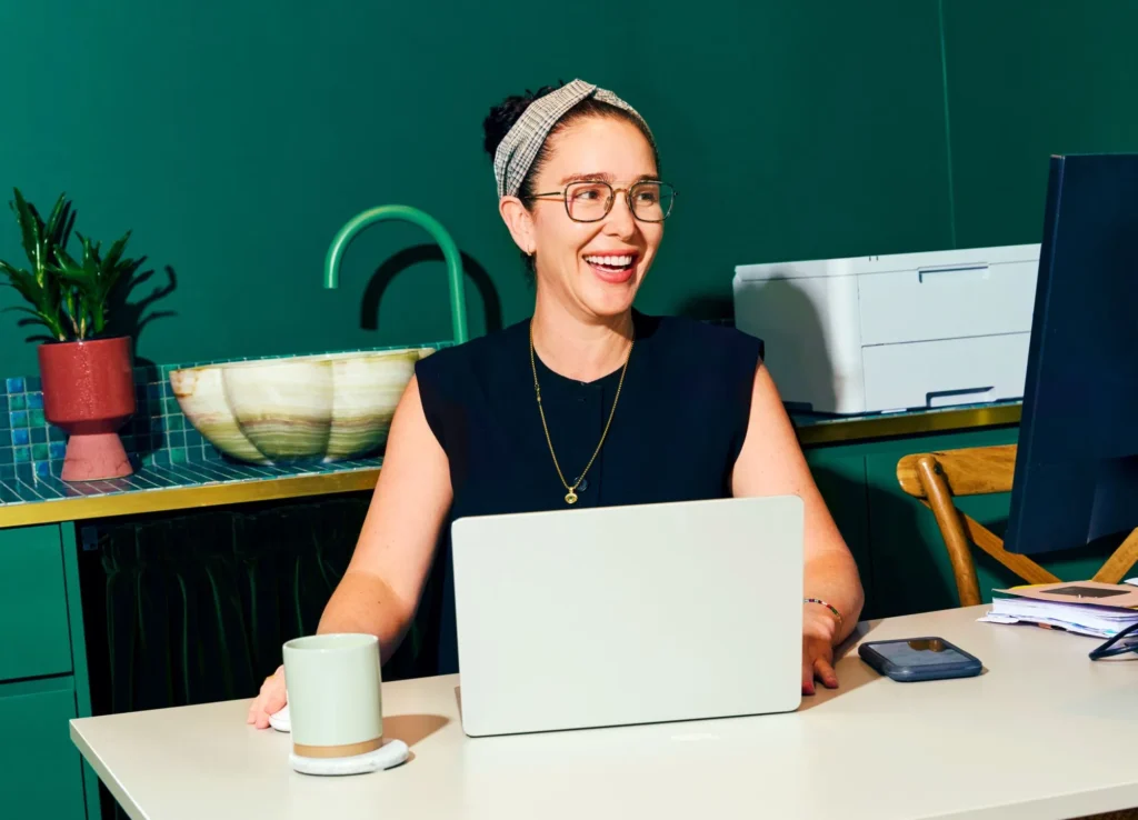 Smiling woman working at a desk with a laptop, coffee mug, and phone in a modern, colorful office space.