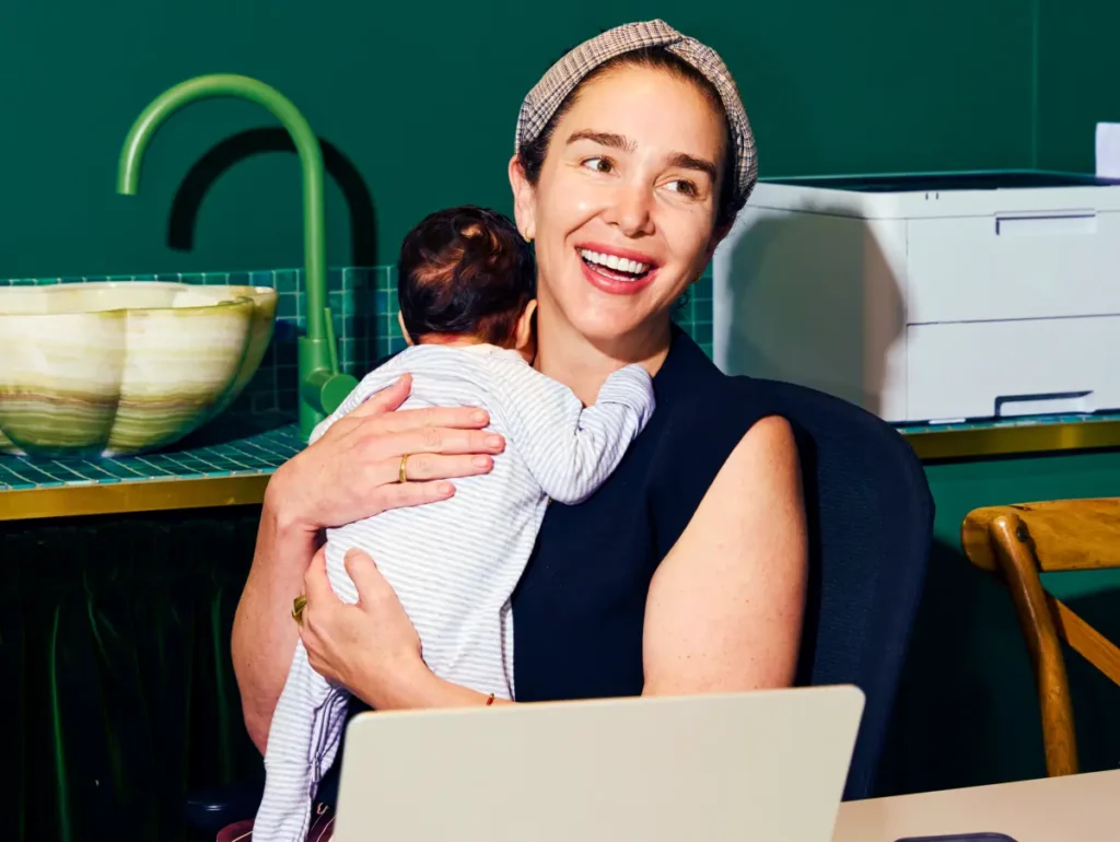 Smiling woman at a desk holding a baby on her shoulder, juggling work and caregiving.