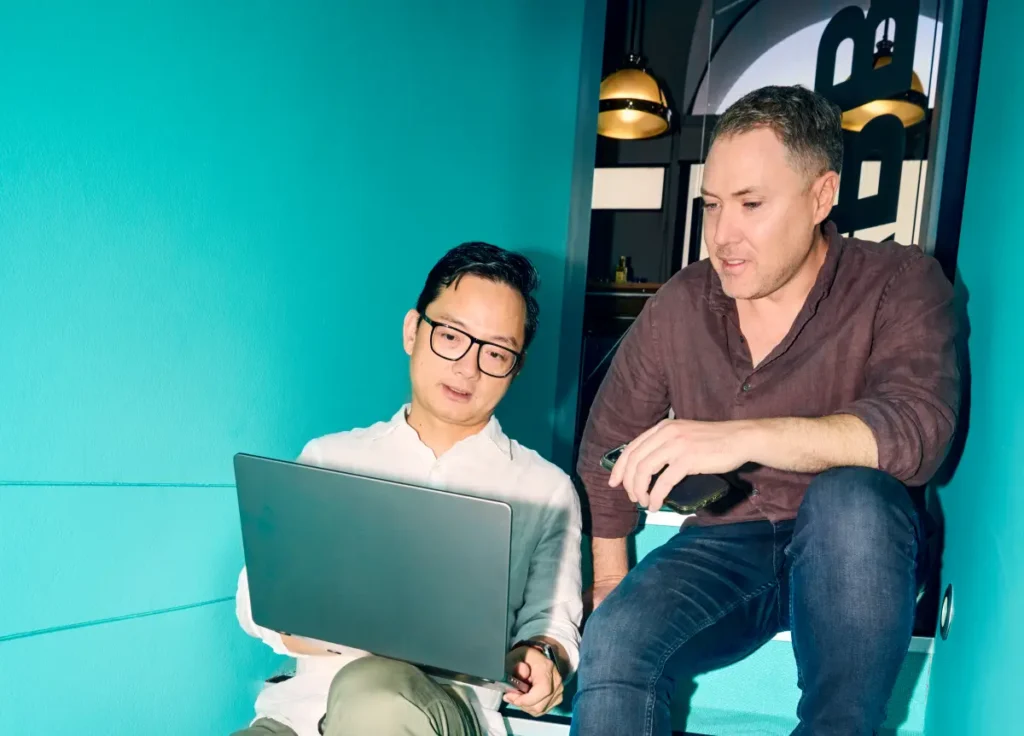 Two men sitting on stairs, looking at a laptop together in a modern office setting.