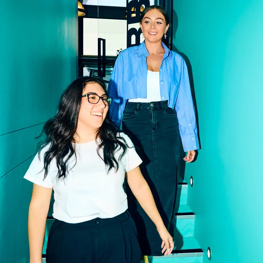 Two women smiling and walking down a turquoise staircase. One wears a white shirt and glasses, the other a blue shirt. Bright, cheerful atmosphere.