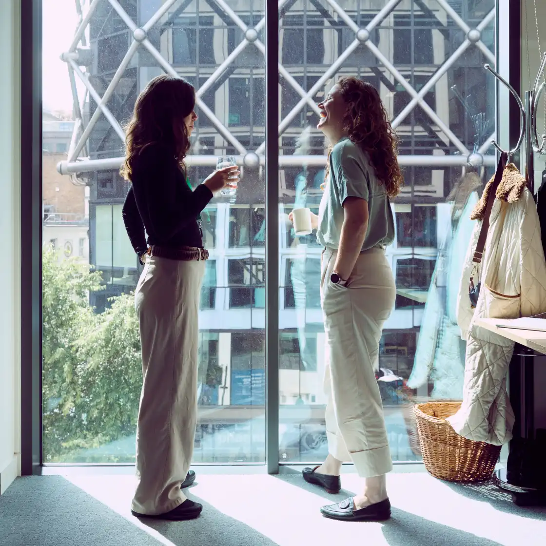 Two women in casual attire stand by a large window, enjoying drinks and laughing. Behind them, a modern building with a geometric design is visible.