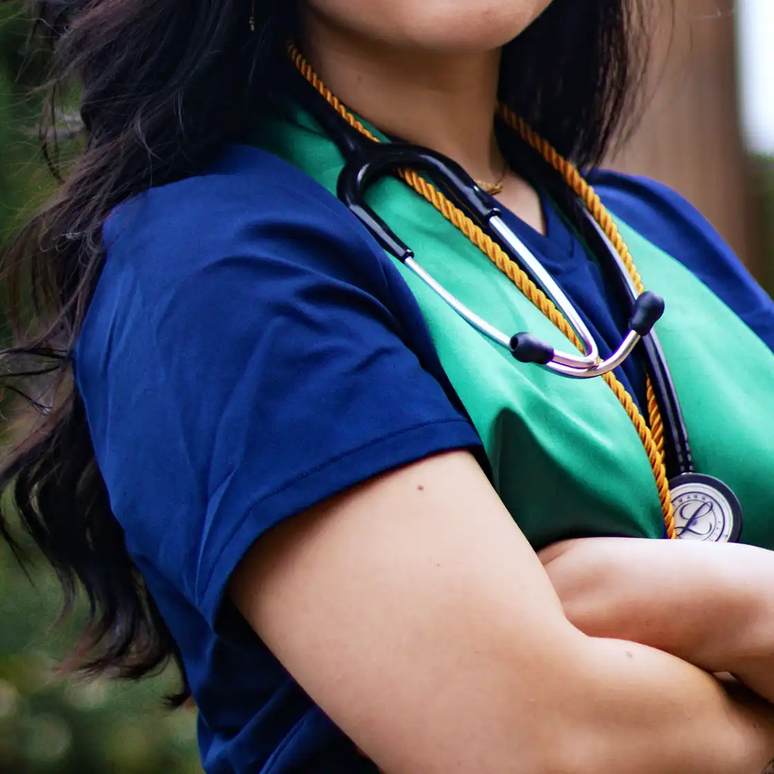 A woman in a blue and green gown poses with arms crossed, wearing a stethoscope and a gold cord, conveying confidence and professionalism.