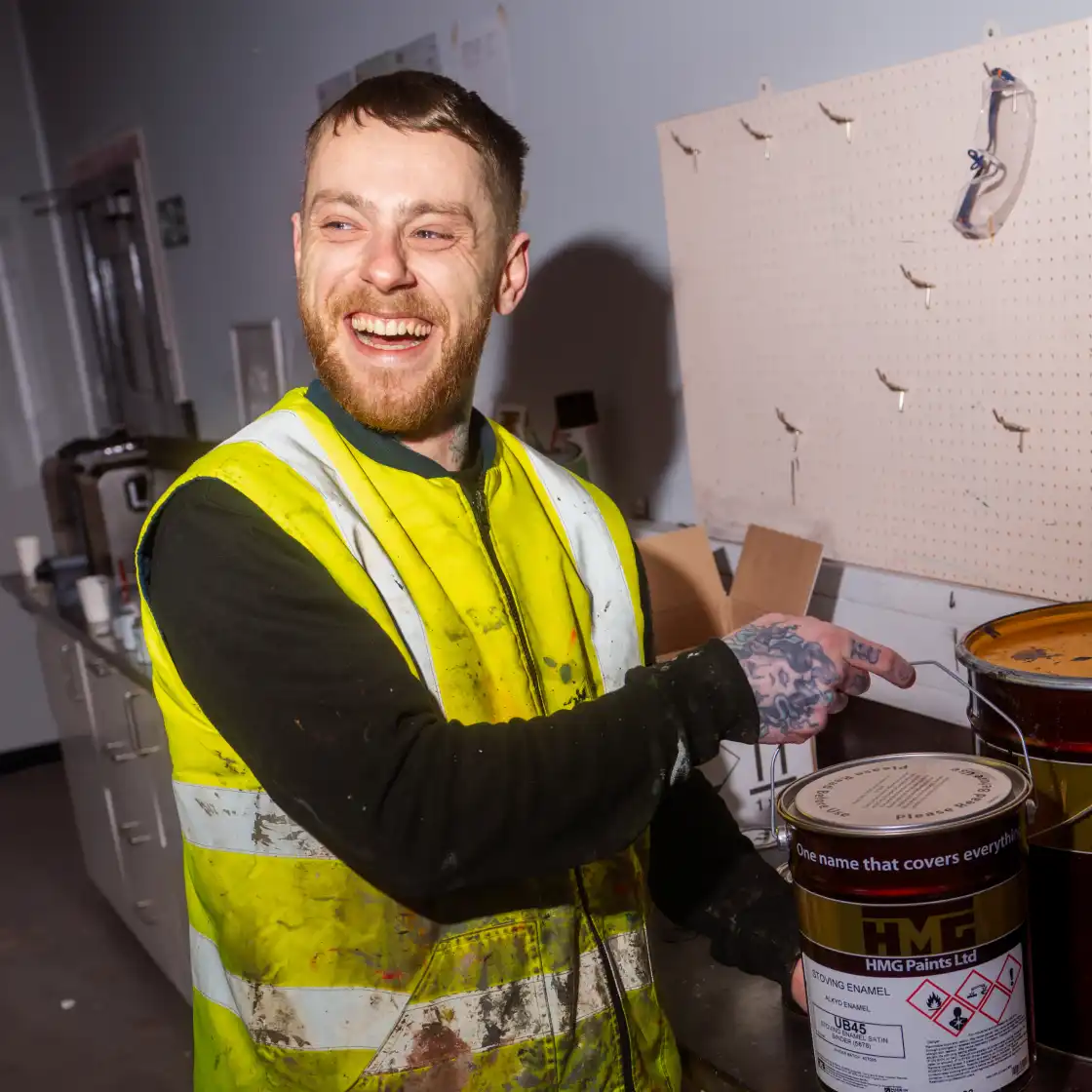 A smiling man in a high-visibility vest points at a paint can in a workshop. The scene is well-lit, with a pegboard and safety goggles in the background.