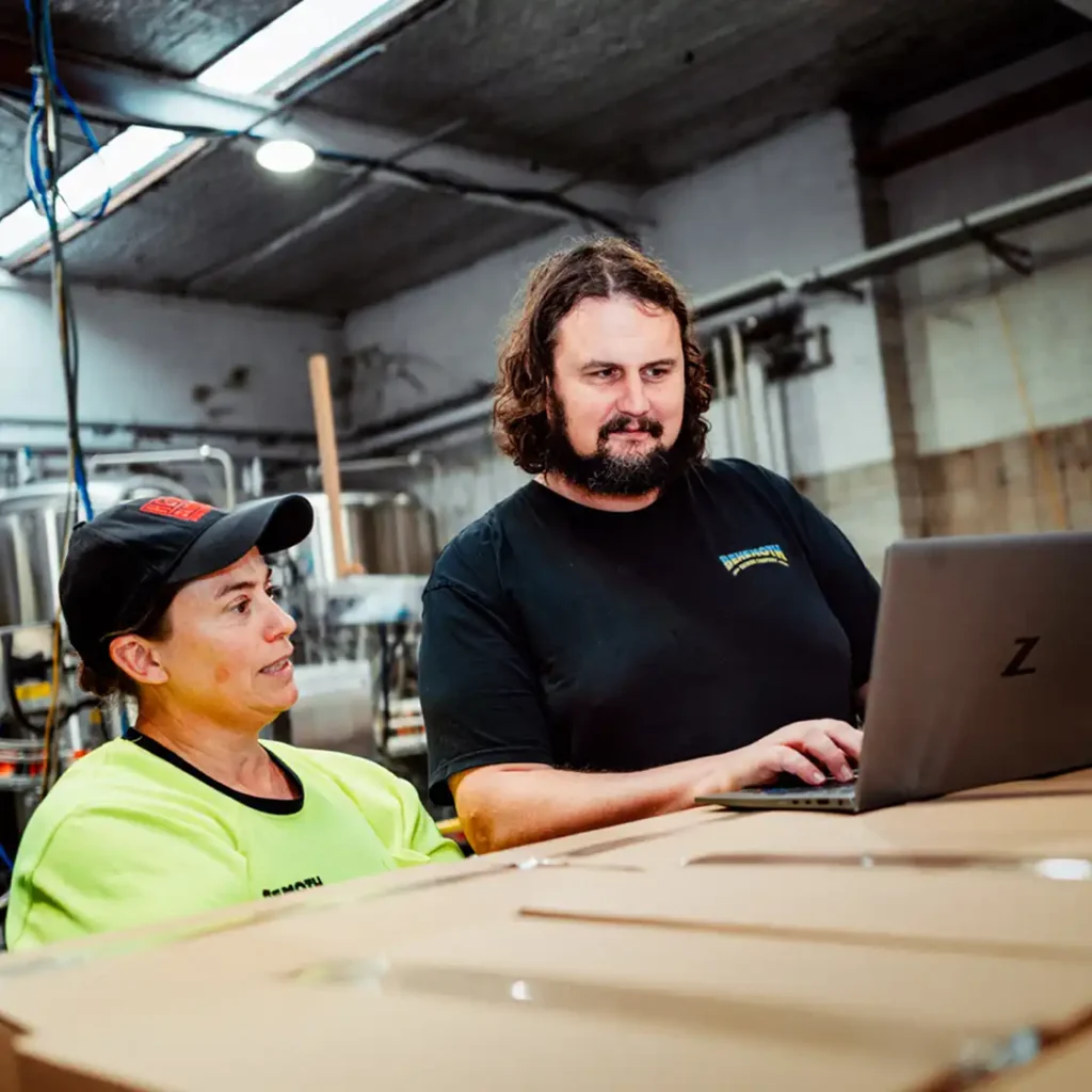 Two people collaborate in an industrial setting. One in a yellow uniform and cap, and another with a beard use a laptop, conveying teamwork and focus.