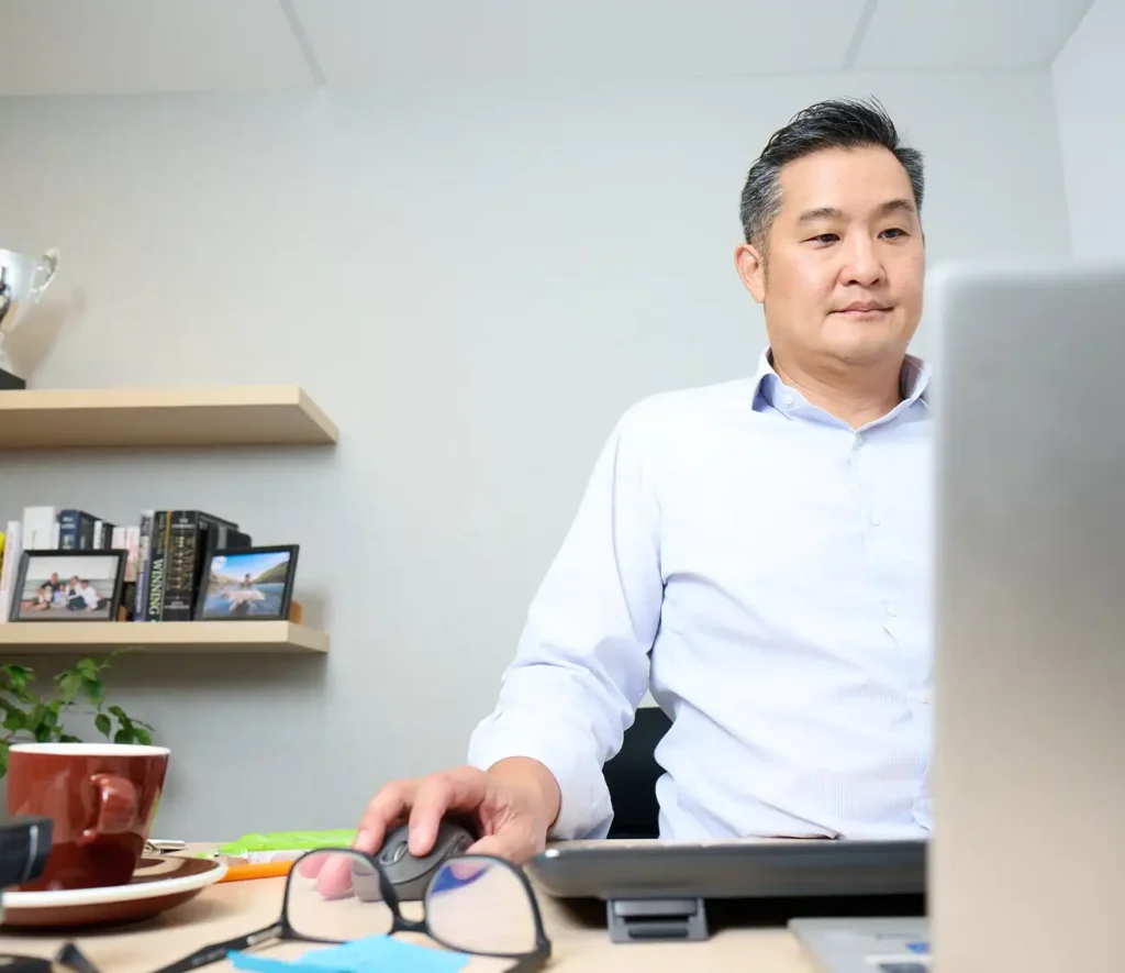 A man sitting at a desk, focused on his laptop computer, with a notepad and pen nearby.