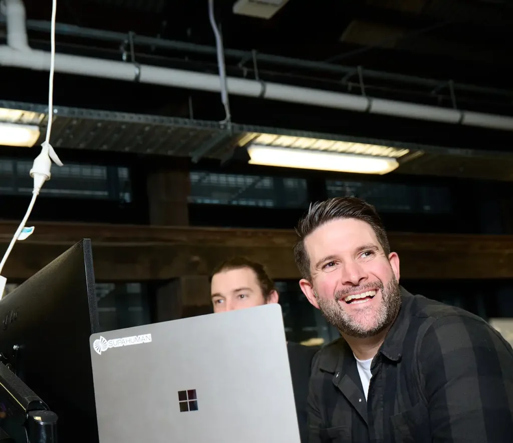 A man smiling while sitting at a computer, engaged and looking content in a well-lit workspace.