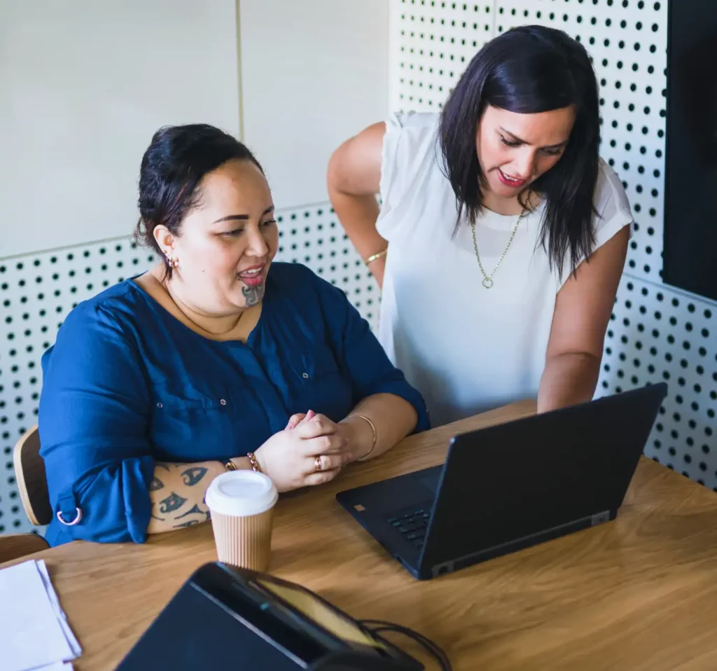 Two women collaborate at a wooden table with a laptop. One points at the screen, while the other listens, smiling. The setting is a modern office.