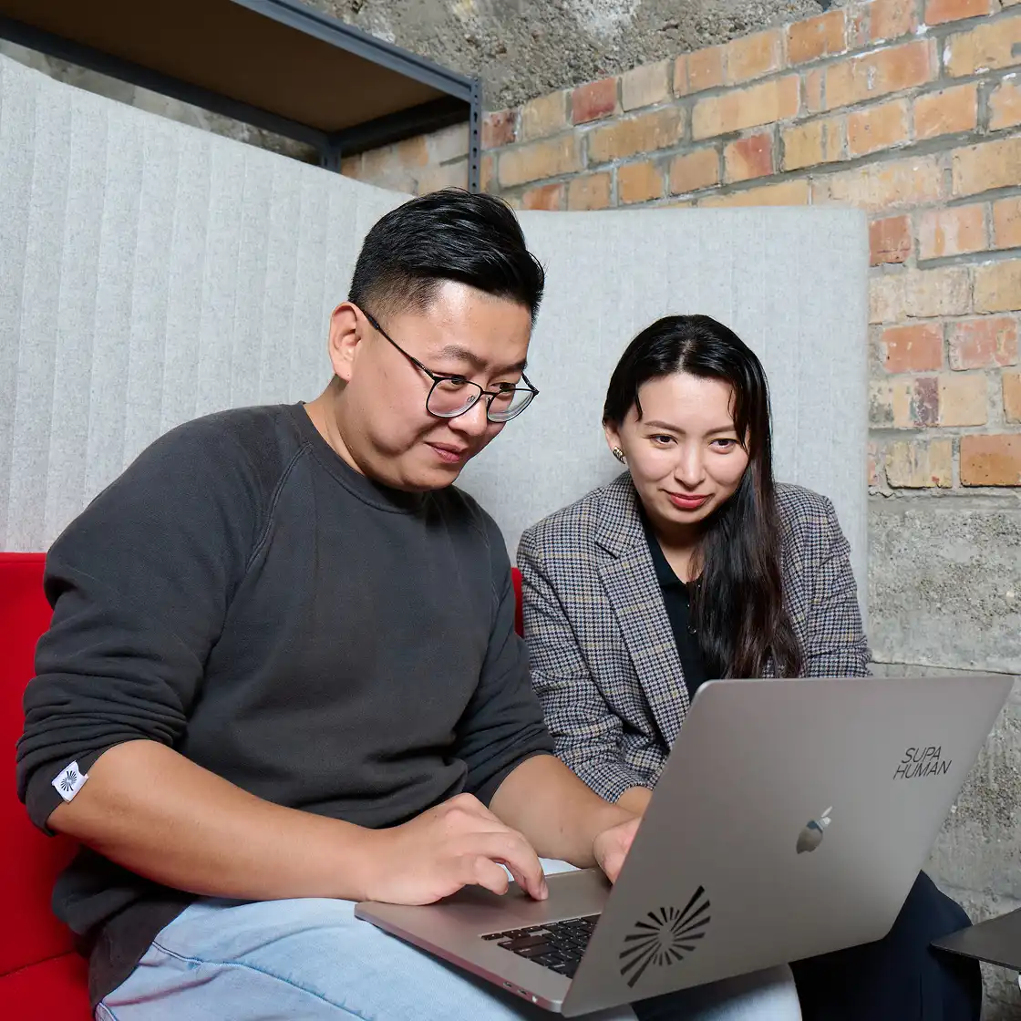 Two people sit closely on a red couch, focused on a laptop screen. The setting has a brick wall and a cozy, collaborative atmosphere.