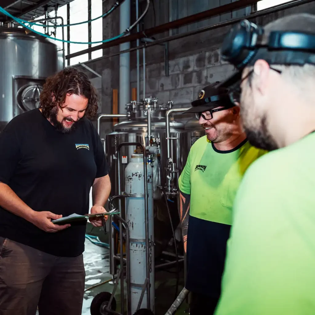 Three men smiling in a brewery setting with steel tanks in the background. One holds a clipboard, fostering teamwork and a positive atmosphere.