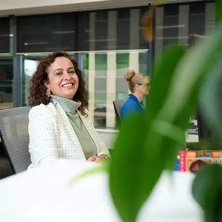 Woman with curly hair in a white jacket smiles while seated at an office desk. Another person works in the background. Green plant in the foreground.