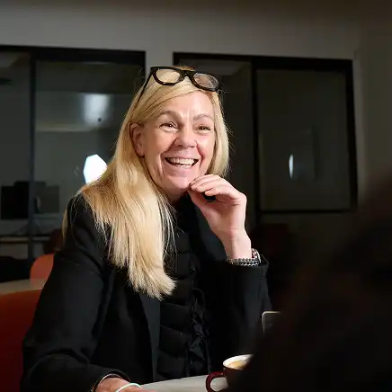 A woman with long blonde hair, wearing glasses on her head and a black jacket, smiles warmly in a well-lit office setting, conveying a friendly tone.