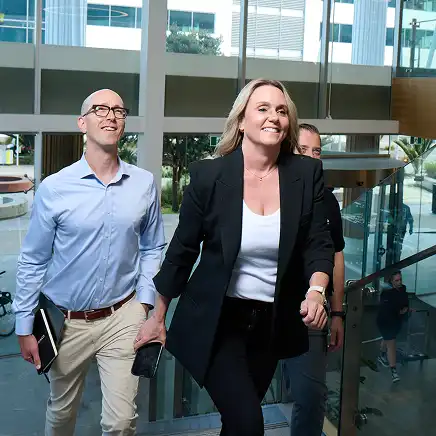 A woman in a black blazer confidently leads two men up a staircase in a modern, bright office building, conveying a positive, professional atmosphere.
