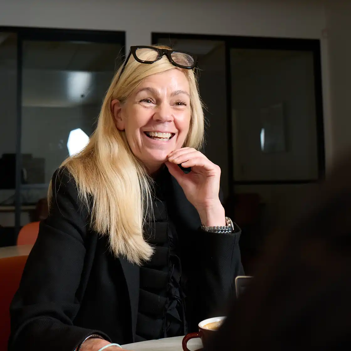 A smiling woman with long blonde hair and glasses on her head sits at a table, engaged in conversation. She wears a black jacket. The atmosphere is warm and friendly.