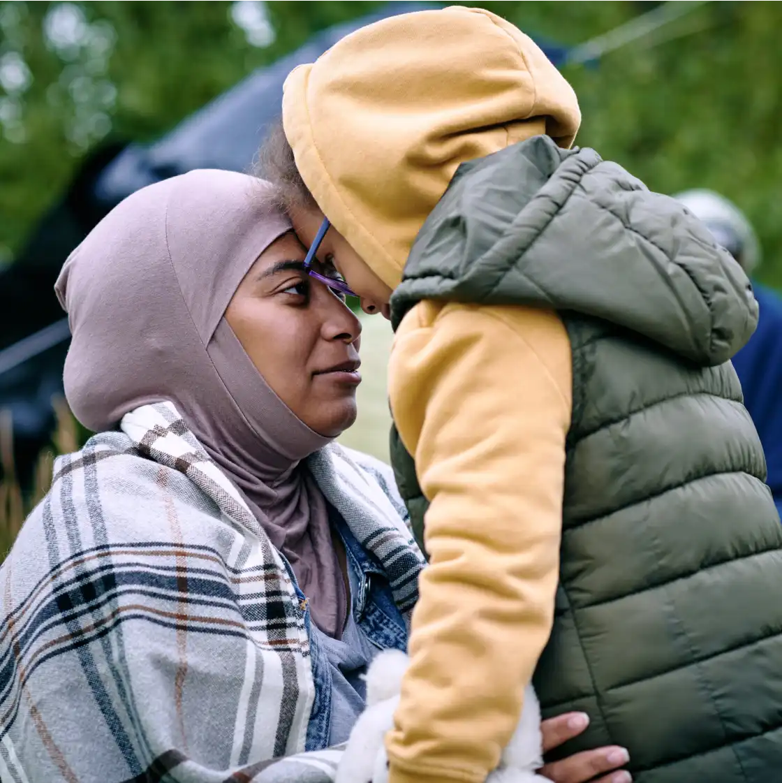 A woman in a hijab and plaid coat lovingly touches foreheads with a child in a yellow hoodie and green vest. The background is a blurred green.