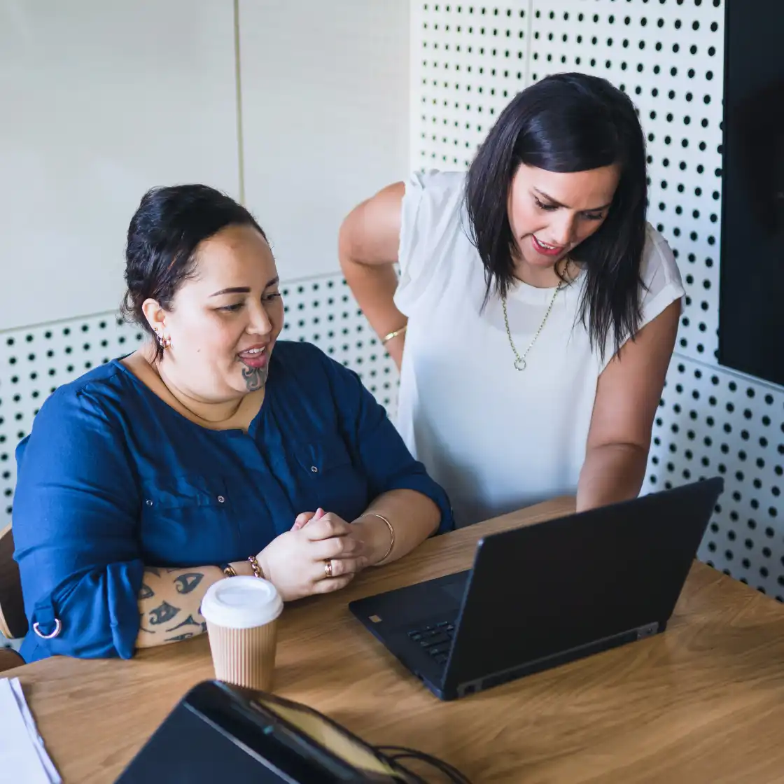 Two women in an office setting, collaborating at a wooden table with a laptop. One in a blue shirt smiles, while the other in white explains. Warm, focused mood.