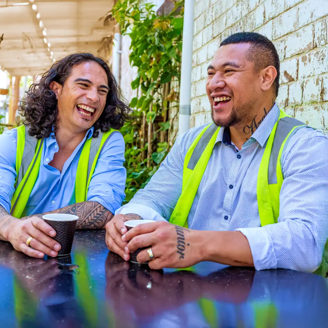 Two men wearing safety vests are sitting and laughing outdoors, each holding a coffee cup. The scene is bright, with a relaxed, joyful atmosphere.