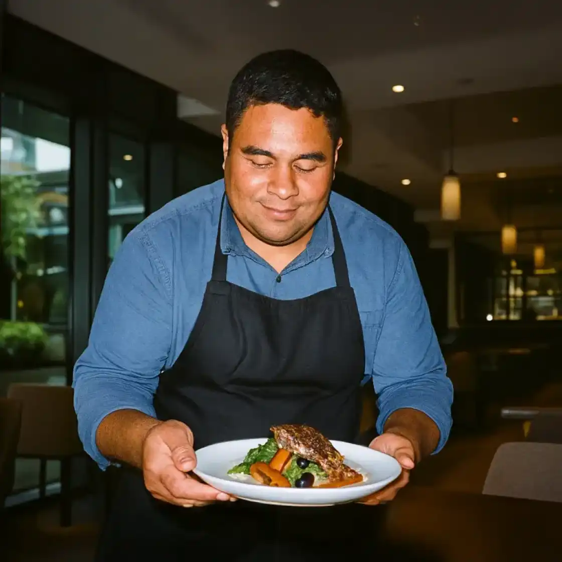 A smiling chef in a blue shirt and black apron presents a plated dish with vegetables and a piece of meat in a warmly lit restaurant setting.
