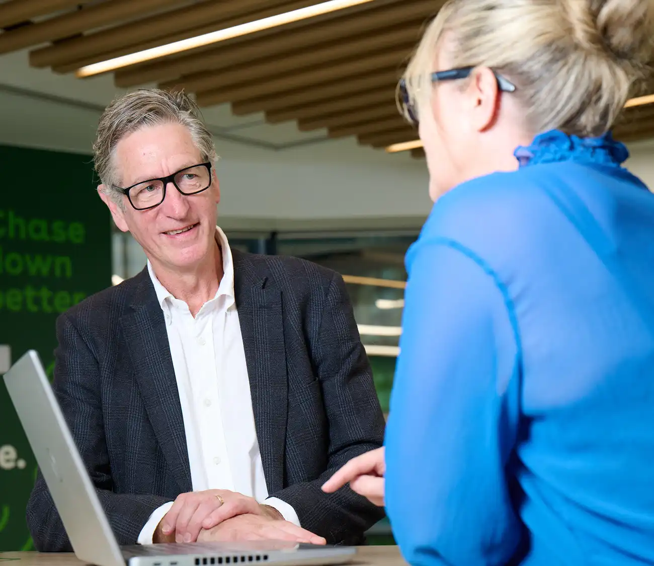 A man in a suit and glasses smiles while talking to a woman in a bright blue blouse. They are sitting at a table with a laptop, discussing in a modern office setting.