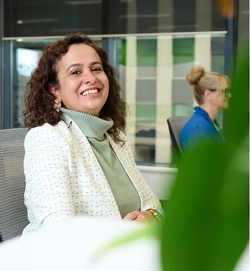 A woman in a light-colored blazer smiles warmly while seated in a modern office. A blurred plant is in the foreground, adding a fresh feel.