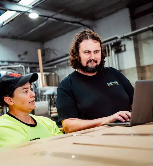 Two people in a warehouse setting work together on a laptop. One, wearing a black shirt, types while the other in a neon shirt watches attentively.