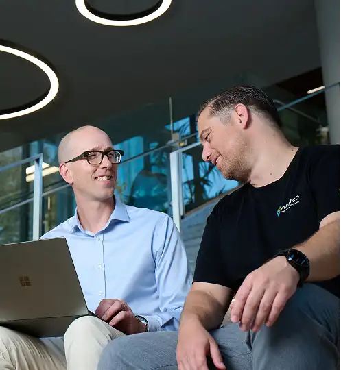 Two men are sitting on steps in a modern office space, smiling and talking. One holds a laptop. The atmosphere is friendly and collaborative.