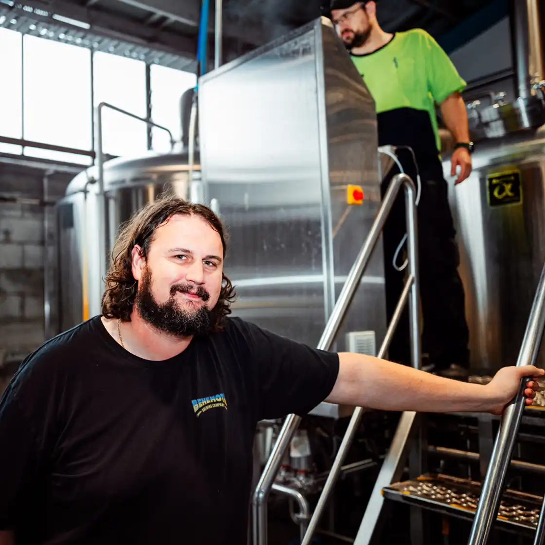 A bearded man smiles at the camera in a brewery, wearing a black shirt. Behind him, a worker in a green shirt stands near a large stainless steel vat. The atmosphere is industrial and friendly.