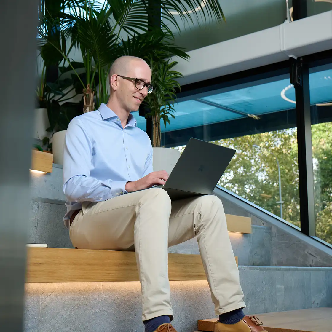 A man in a light blue shirt and beige pants sits on wooden steps with a laptop, smiling. Bright, modern indoor space with large plants and glass windows.